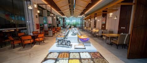 A buffet setup with various food items in the foreground, a long dining area with tables and chairs, and a modern ceiling design.