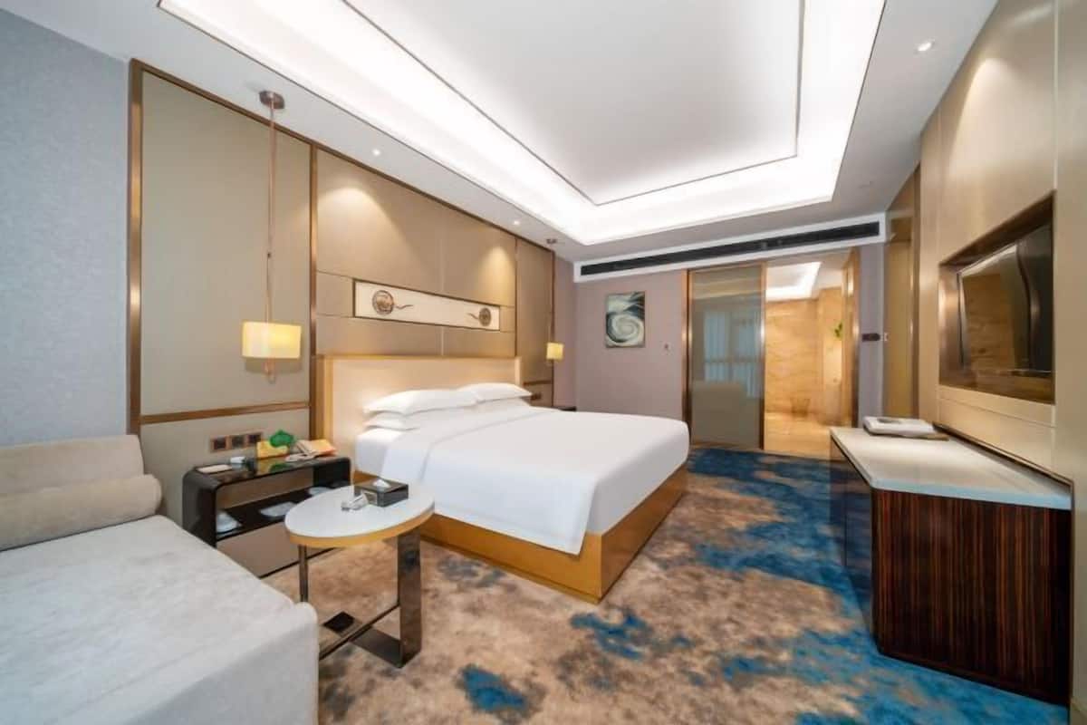 A modern hotel room with a large bed, a desk, a chair, a TV, and a bathroom visible through a glass door.