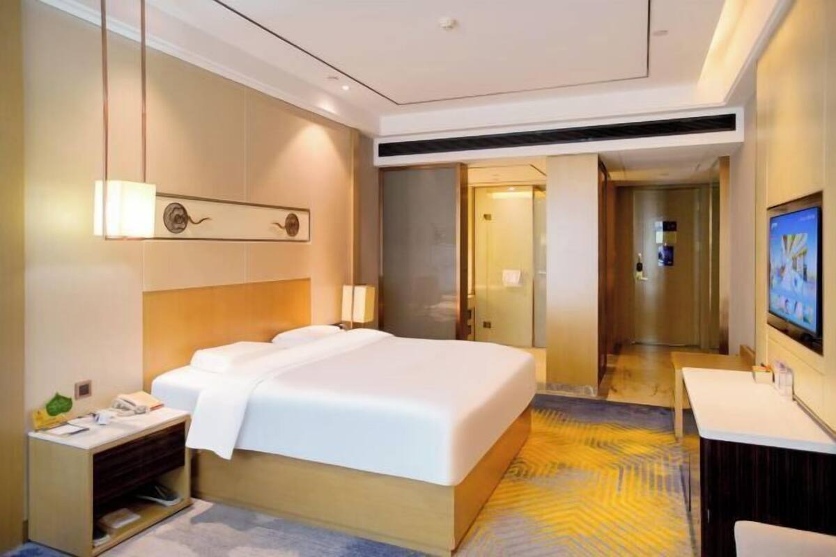 A hotel room with a large bed, bedside table, lamp, TV, and a view of the hallway.