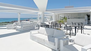 A modern outdoor lounge area with white furniture, including sofas and tables, overlooking a coastal cityscape.