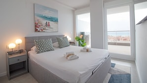 A modern bedroom with a large bed, bedside lamps, a painting, and a view of the city through a balcony.