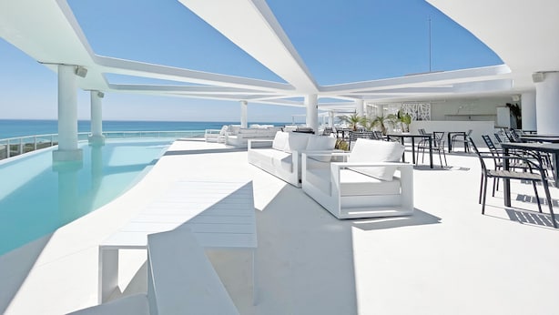 A modern outdoor lounge area with a pool, white furniture, and a clear view of the ocean.