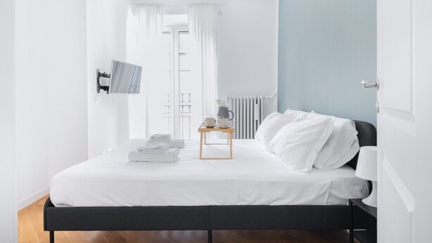 A modern bedroom with a bed, a bedside table with a lamp, a TV mounted on the wall, and a window with curtains.