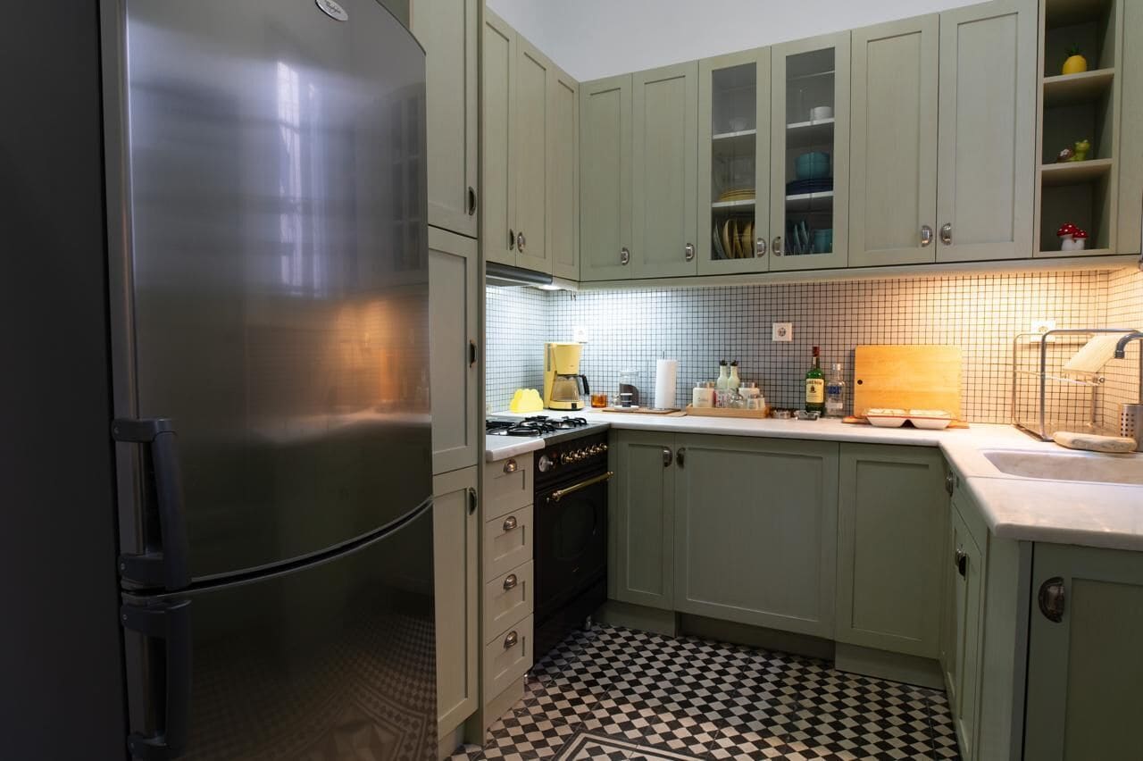 A kitchen with light green cabinets, a stainless steel refrigerator, a tiled backsplash, and a checkered floor.