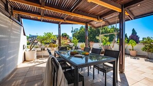 Un patio techado con una mesa de vidrio y sillas de ratán negro, rodeado de plantas en macetas y vistas a la ciudad.
