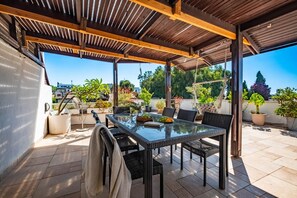 Una veranda riparata con un tavolo in vetro e sedie in vimini nero, circondata da piante in vaso e vista sulla città.