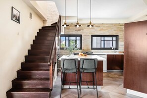 A kitchen with a central island, dark wooden staircase, and hanging pendant lights.