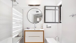 Un baño moderno con cabina de ducha de vidrio, un espejo redondo, un lavabo blanco con gabinete de madera, un inodoro y un perchero para toallas.