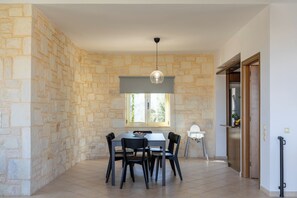 A dining area with a stone wall, a table with chairs, a hanging light fixture, and a window with a view of greenery. - Sense Of Dream Villas (Platanias)