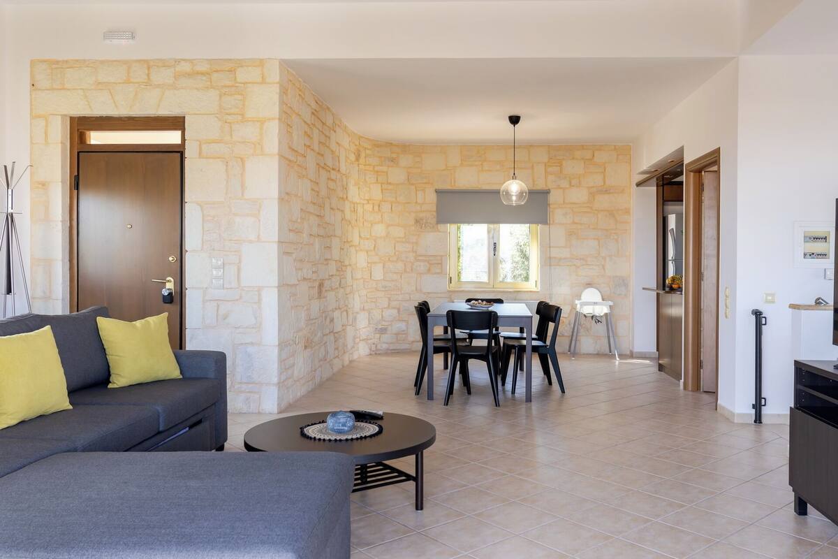 A modern living room with a stone wall, a grey sofa with yellow cushions, a dining area with a table and chairs, and a kitchen area with a fridge and cabinets.