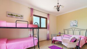 Quarto com beliches, uma cama de solteiro, uma cadeira, janela com cortinas, um lustre e uma planta na parede.