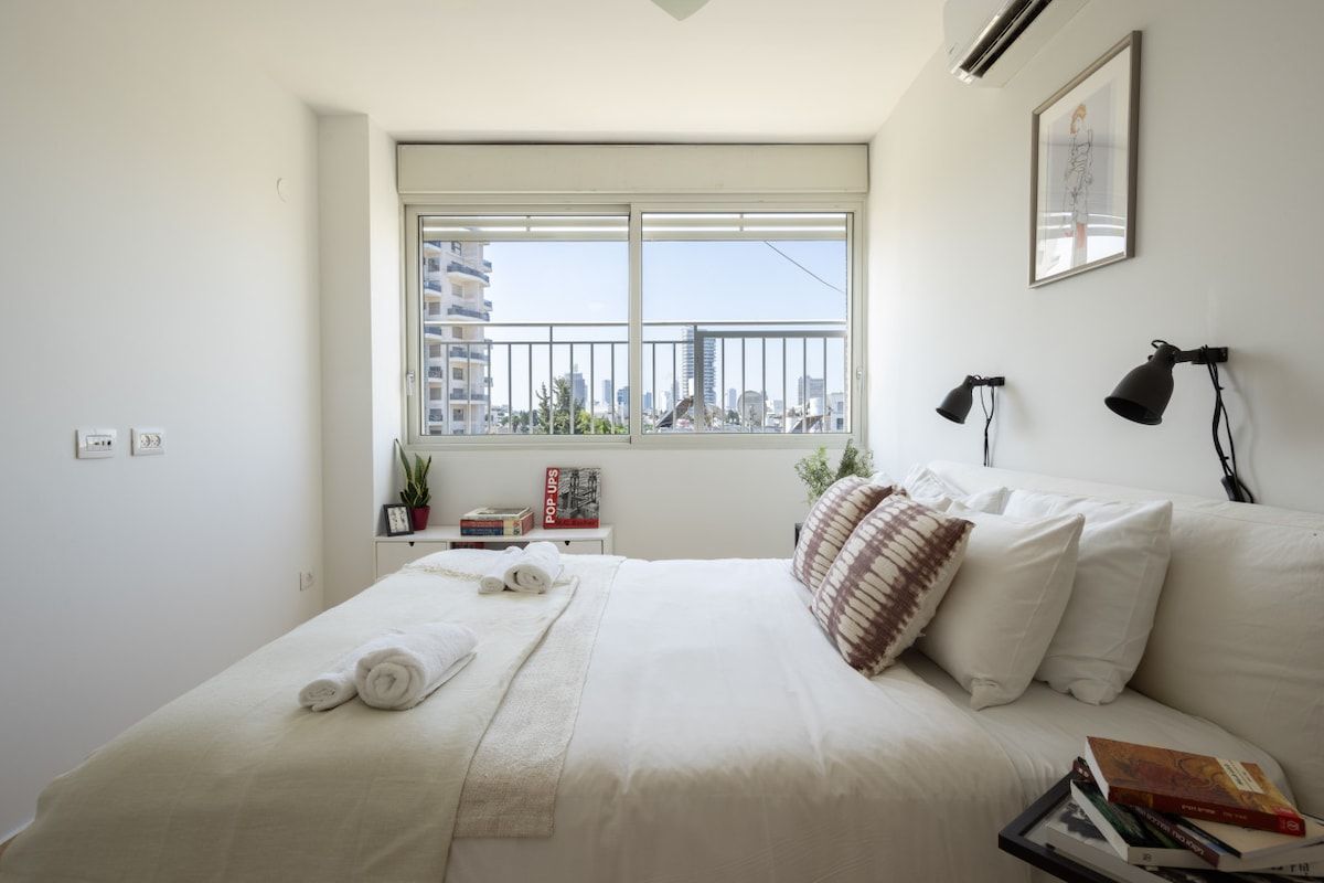 A bedroom with a large bed, a window with a city view, a bedside table with books and a plant, and a framed picture on the wall.