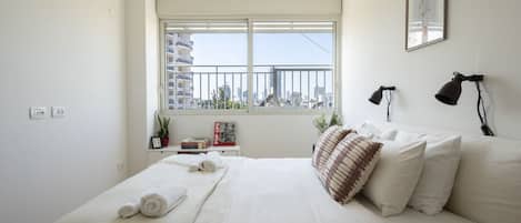 A bedroom with a large bed, a window with a city view, a bedside table with books and a plant, and a framed picture on the wall.