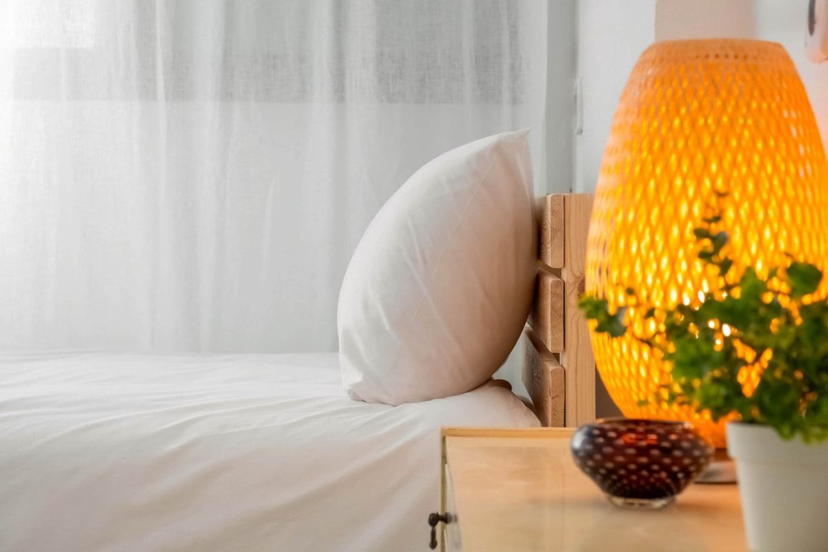 Ein Bett mit weißer Bettwäsche, ein Nachttisch aus Holz mit einer Lampe, einer Schale und einem Blumentopf.