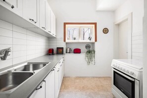 Une cuisine compacte avec des meubles blancs, des éviers en acier inoxydable, une bouilloire rouge et une petite fenêtre au-dessus du plan de travail.
