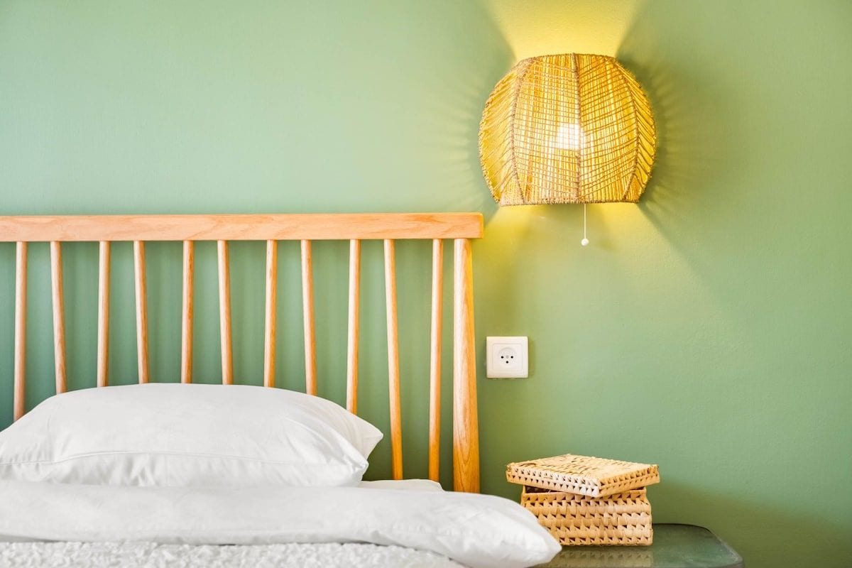A bed with a wooden headboard, a white pillow, and a white blanket. There is a woven lamp on a wall to the right of the bed.