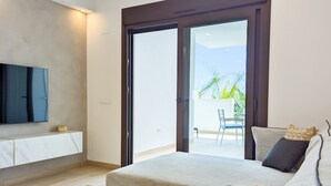 Un dormitorio moderno con una cama grande, un televisor montado sobre un mueble de mármol y un ventanal corredizo que da a un balcón con mobiliario exterior.