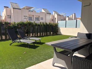 A patio area with a table and two lounge chairs, surrounded by artificial grass and a hedge.