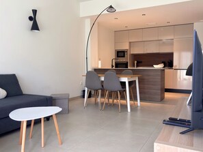 En moderne stue med en sofa, et lille bord og et spiseområde med stole. Et køkken er synligt i baggrunden med en mikrobølgeovn og ovn.