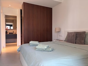 Un dormitorio moderno con una cama grande, un panel de pared de madera, una mesita de noche con lámpara y un baño visible a través de una puerta abierta.