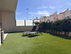 Una terraza en la azotea con césped artificial, dos sillones de descanso, un muro blanco y una fila de plantas.