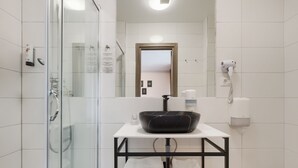 Un baño moderno con cabina de ducha de vidrio, un lavabo negro sobre encimera blanca y un secador de pelo fijado en la pared.