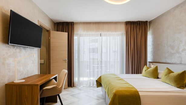 Habitación de hotel con cama, escritorio, silla, televisor y un amplio ventanal con cortinas.