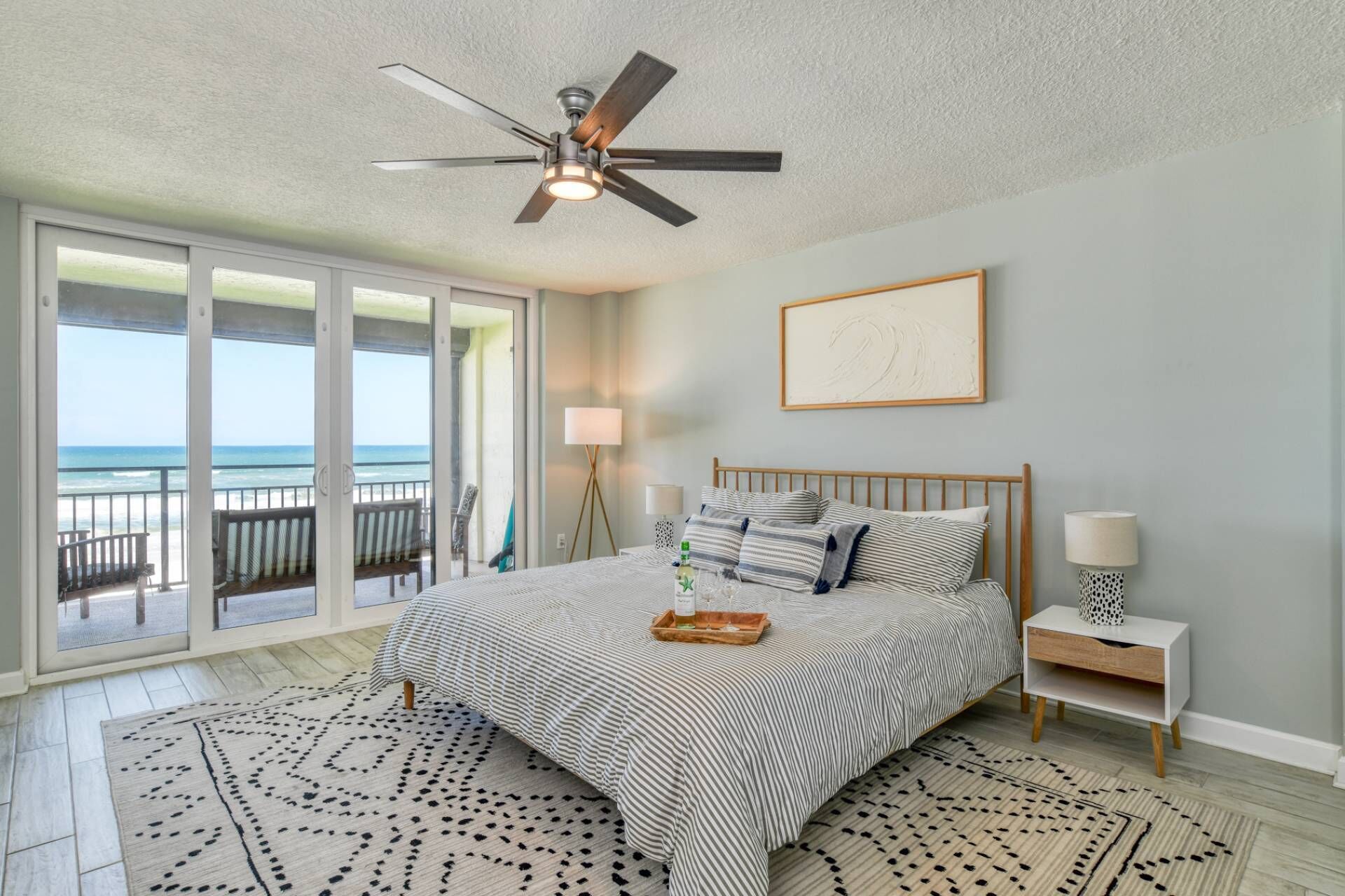 A bedroom with a bed, a ceiling fan, a nightstand, a lamp, and a view of the ocean through large sliding doors.