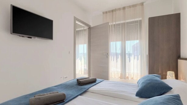 Una habitación de hotel moderna con un televisor de pantalla plana montado en la pared, una cama con ropa de cama azul y una ventana con cortinas traslúcidas.