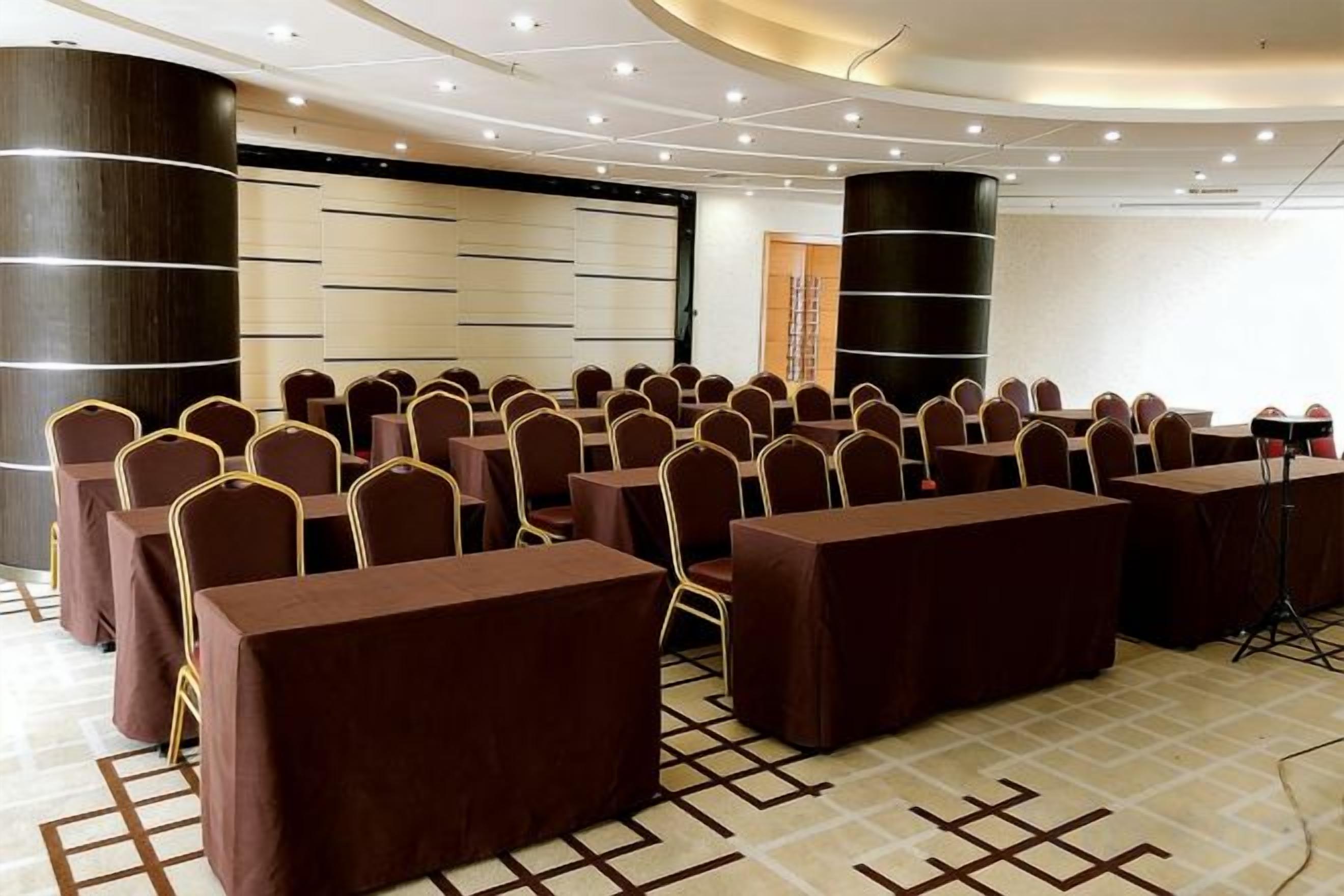 Un salón de conferencias con filas de mesas y sillas, un escenario y una pantalla grande.