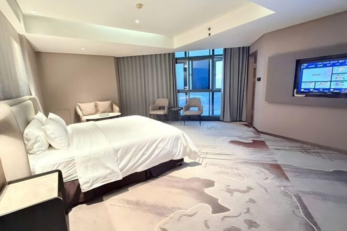 Una habitación de hotel moderna con una cama grande, un área de descanso con dos sillones, un televisor montado en la pared y vistas a la ciudad a través de amplios ventanales.
