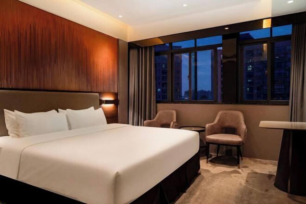 Une chambre d’hôtel avec un grand lit, un bureau, deux chaises et une vue sur la ville au crépuscule.