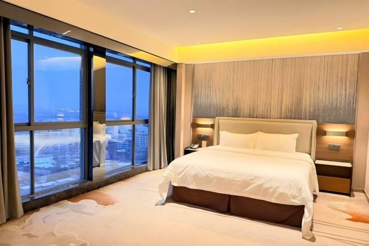 Habitación de hotel moderna con una cama grande, una mesita de noche y vistas de la ciudad a través de ventanales del suelo al techo.
