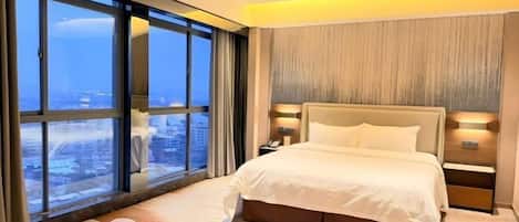 A modern hotel room with a large bed, a nightstand, and a view of the city through floor-to-ceiling windows.