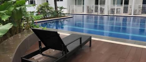 A poolside area with a lounge chair, a pool, and a building with balconies in the background.