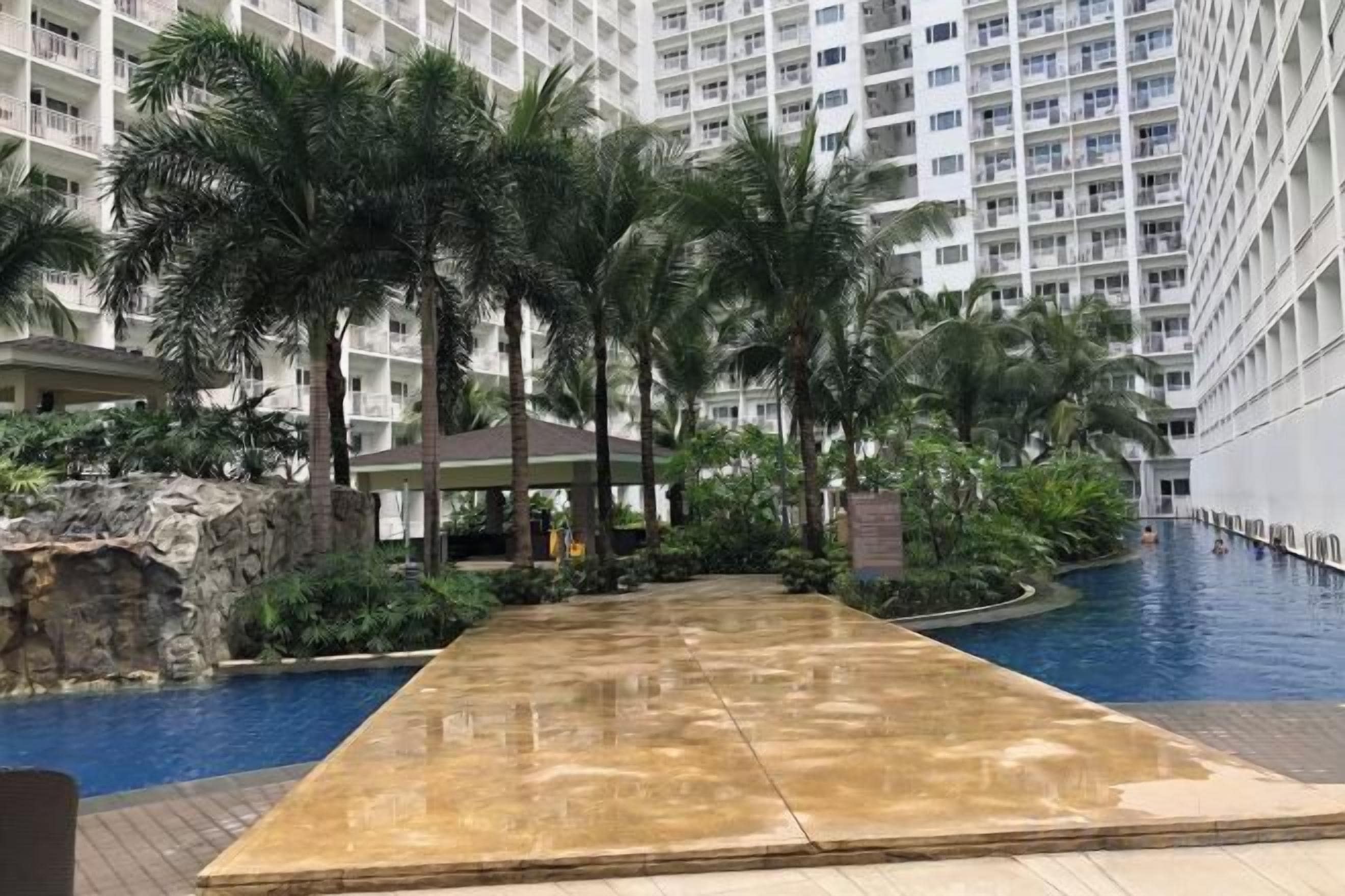 A swimming pool area with a long, narrow pool, surrounded by palm trees and a building with multiple balconies.