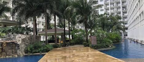 A swimming pool area with a long, narrow pool, surrounded by palm trees and a building with multiple balconies.