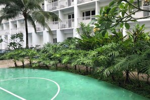Ein grüner Basketballplatz im Freien, umgeben von üppiger Vegetation und einem weißen Gebäude mit Balkonen.