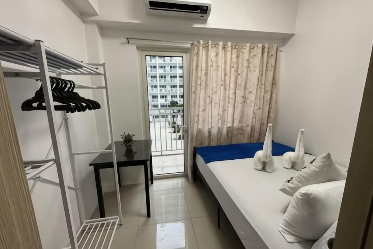 Un dormitorio con cama, una mesita, un ventanal con cortinas y un balcón.