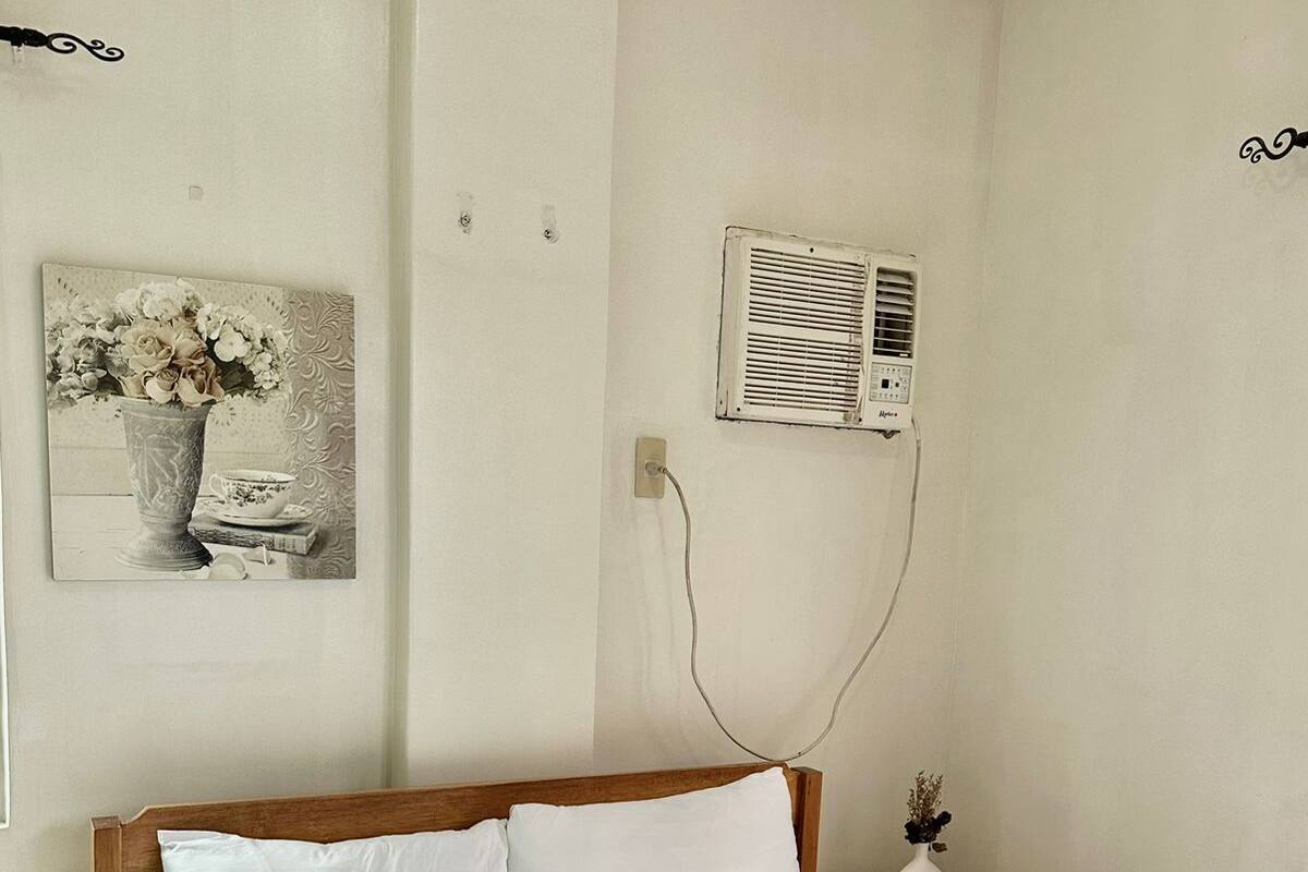 Un lit bien fait, avec du linge de lit blanc, une tête de lit en bois et une petite table de chevet ornée d’un vase et de quelques livres.