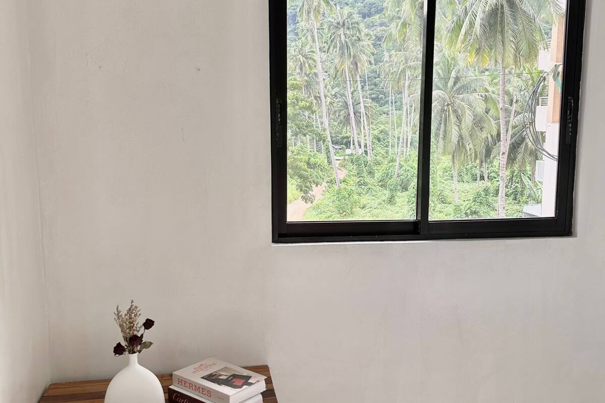 Une pièce avec une fenêtre donnant sur un paysage tropical, une petite table en bois sur laquelle sont posés un vase et un livre, et des murs blancs sobres.