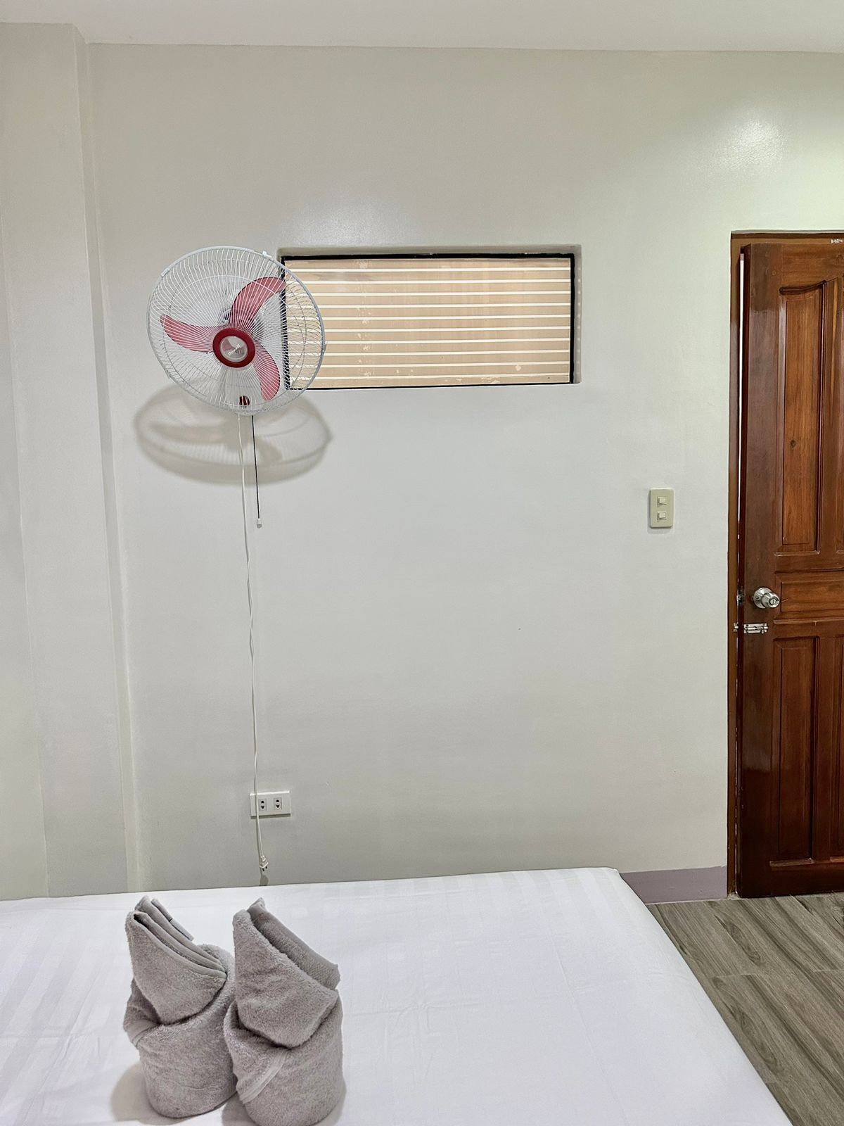 A hotel room with a bed, a standing fan, a window with blinds, and a wooden door.