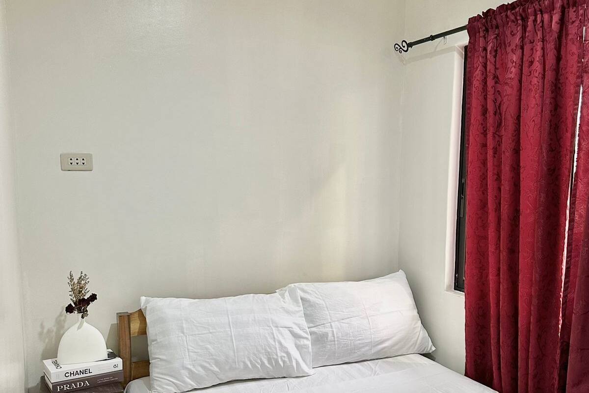 Un lit bien fait, avec du linge de lit blanc, une paire de chaussures posée sur le lit, une table de chevet avec un vase et des livres, et une fenêtre avec des rideaux rouges.