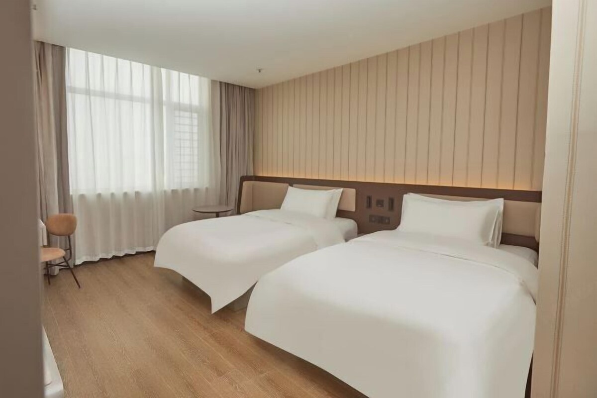Une chambre d’hôtel avec deux lits, une petite table, une chaise et une grande fenêtre avec des rideaux.
