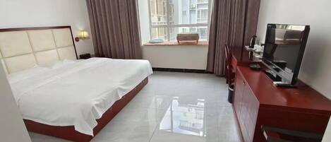 A hotel room with a bed, a TV, a desk, a window with curtains, and a view of buildings outside.
