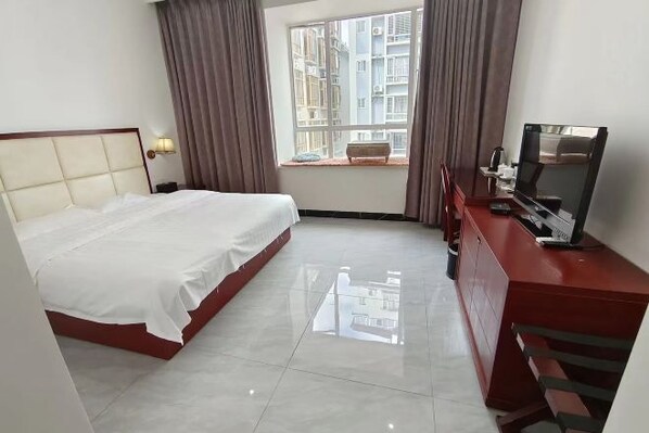 A hotel room with a bed, a TV, a desk, a window with curtains, and a view of buildings outside.