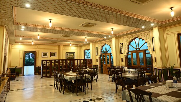 A spacious dining area with multiple tables and chairs, ornate ceiling design, and large windows with decorative glass panes. - Hotel Rime Vista (Jaipur)