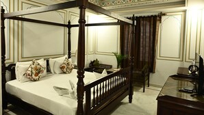A four-poster bed with a wooden frame, white bedding, and floral pillows. There is a desk with a computer and a chair in the room. - Hotel Rime Vista (Jaipur)
