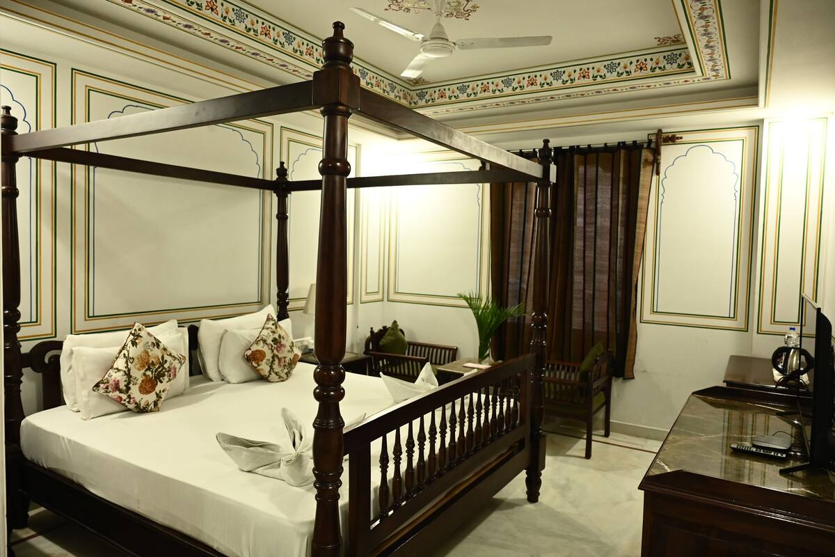 Una cama con dosel de madera, ropa de cama blanca y almohadas con motivos florales. Hay un escritorio con una computadora y una silla en la habitación.