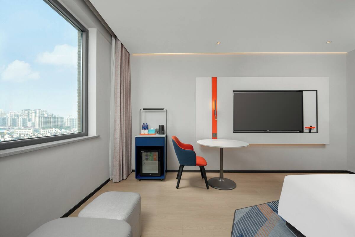 A modern hotel room with a large window, a flat-screen TV mounted on the wall, a small table, a blue and orange chair, a minibar, and a bed.
